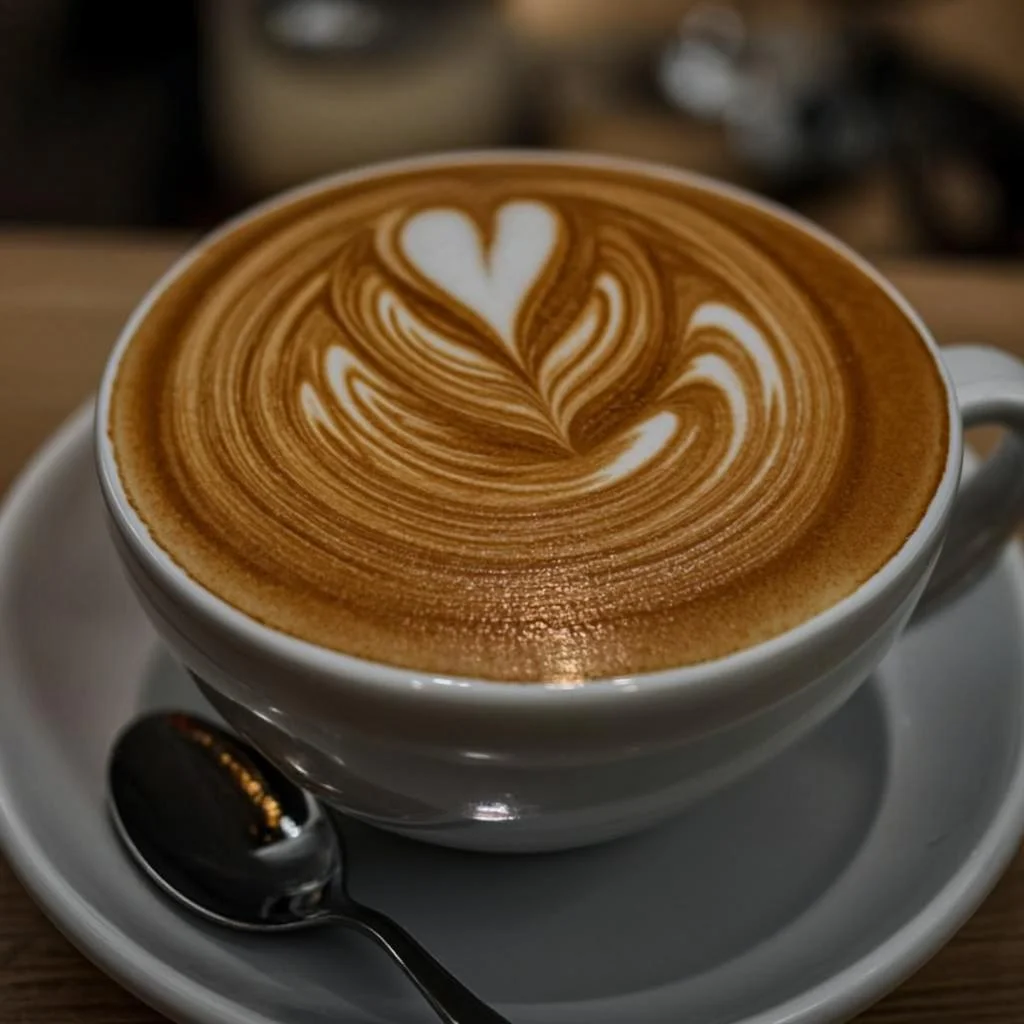 A close-up, high-quality photograph of a creamy latte in a white ceramic cup, featuring beautiful detailed heart-shaped and leaf-style latte art on the coffee surface. The cup sits on a matching saucer with a dark spoon beside it. The warm brown tones, soft lighting, shallow depth of field, and blurred café background create a cozy, elegant, premium coffee-shop atmosphere.